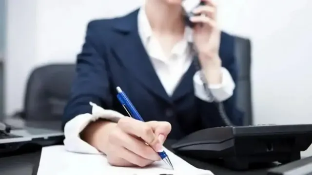 O persoană purtând sacou și cămașă scrie pe o foaie de hârtie cu un pix albastru, în timp ce ține un telefon la ureche.