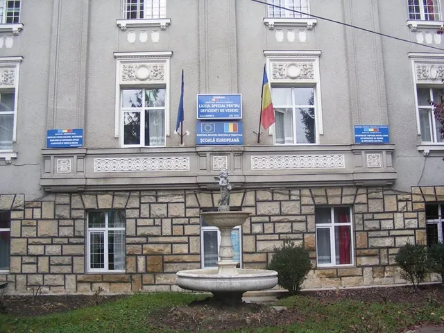O clădire cu fațadă gri și bază din piatră, având ferestre albe, un balcon lung și un fântâna în fața acesteia.