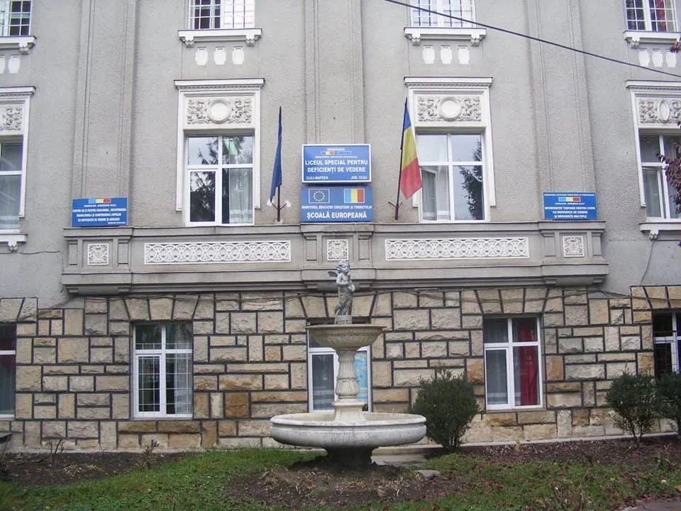 O clădire cu fațadă gri și bază din piatră, având ferestre albe, un balcon lung și un fântâna în fața acesteia.