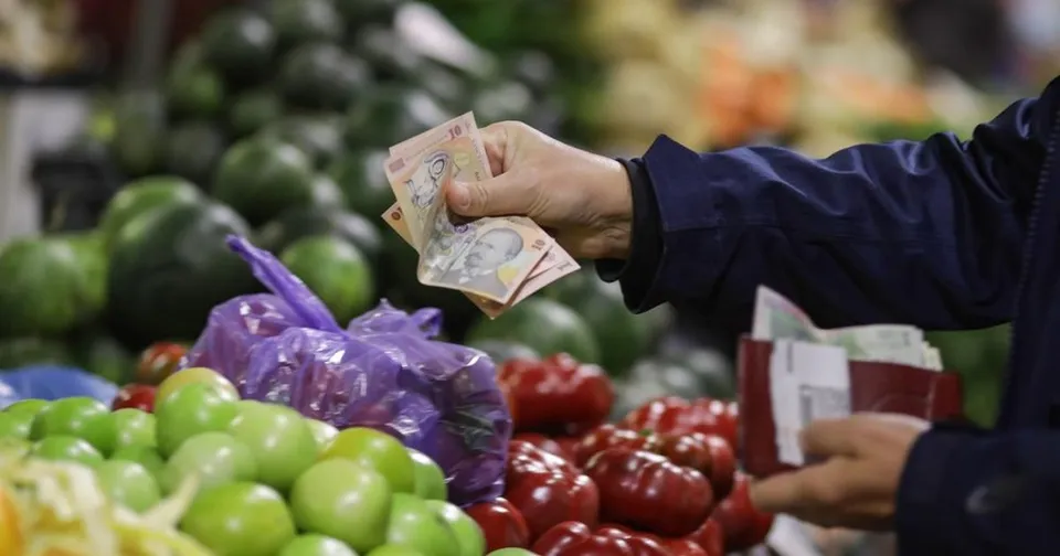 O persoană ține un portofel și oferă câteva bancnote deasupra unor legume proaspete dintr-un magazin.
