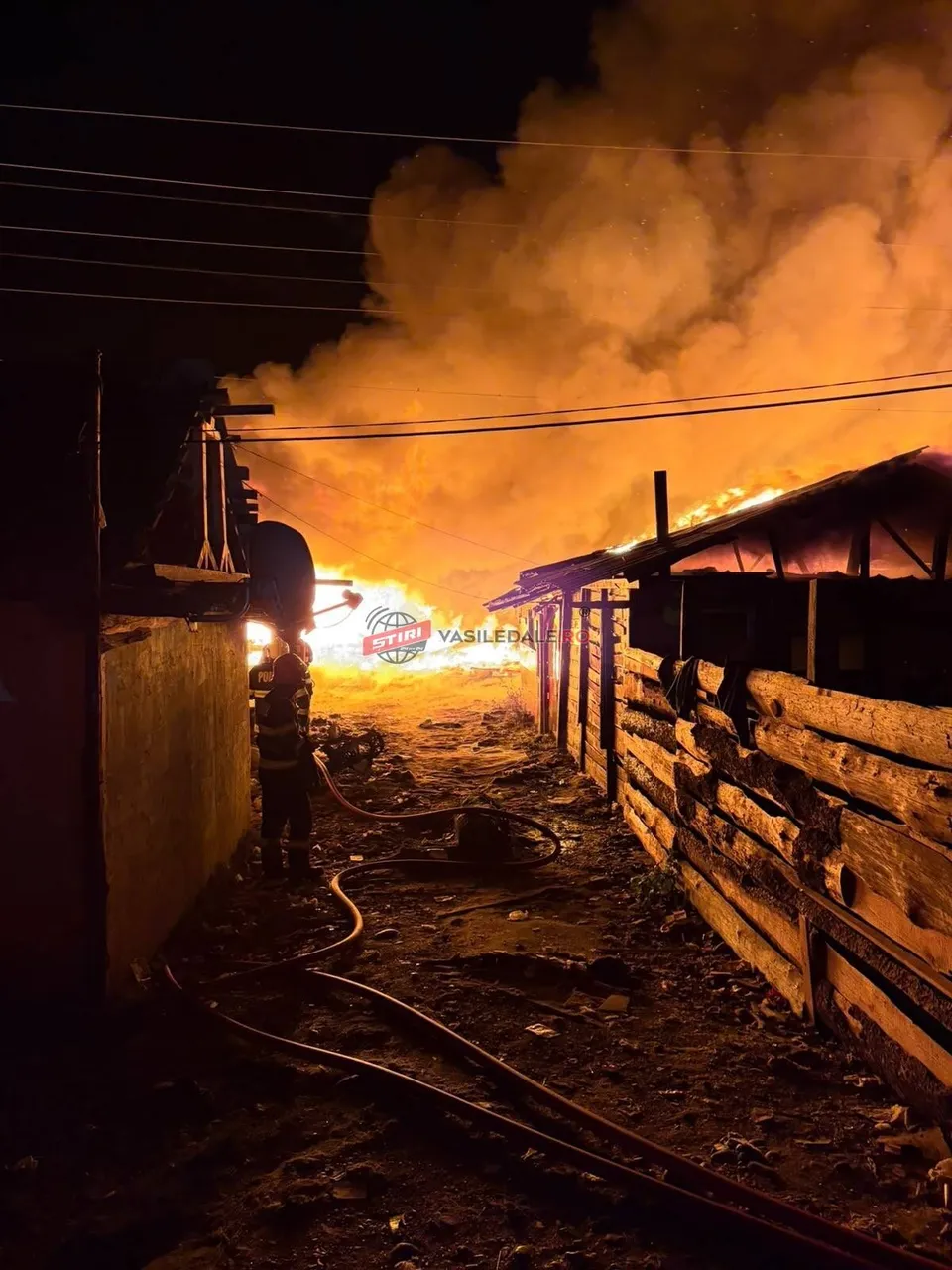 Un pompier stă lângă o structură de lemn în timp ce flăcări și fum dens se ridică din spatele unor cabluri electrice.