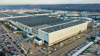O vedere aeriană prezintă un complex industrial extins cu clădiri mari de culoare deschisă, parcuri de mașini și drumuri cu vehicule circulând în apropiere.