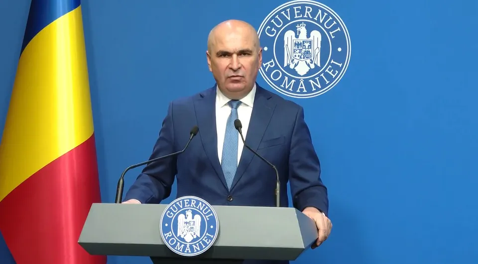 Un bărbat cu părul scurt și deschis la culoare poartă un costum albastru, cămașă albă și cravată albastră, stând în spatele unui podium cu un emblema oficială.