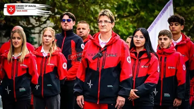 Un grup de persoane poartă haine uniforme de culoare roșie și neagră cu embleme albe, stând în rând în fața unor copaci verzi.