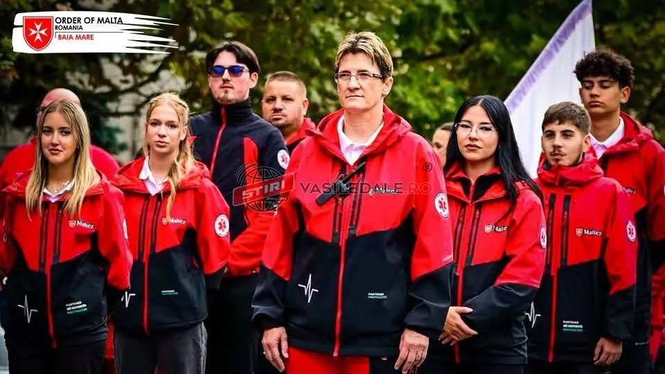Un grup de persoane poartă haine uniforme de culoare roșie și neagră cu embleme albe, stând în rând în fața unor copaci verzi.
