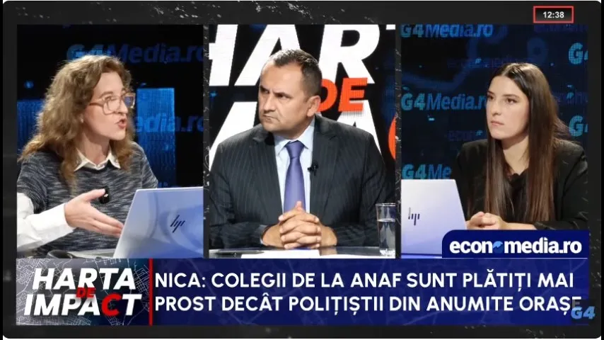 Trei persoane stau la o masă într-un studio de televiziune, având în față laptopuri și un pahar cu apă.