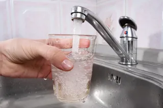 O mână ține un pahar de sticlă sub un robinet metalic din care curge apă într-o chiuvetă de inox.