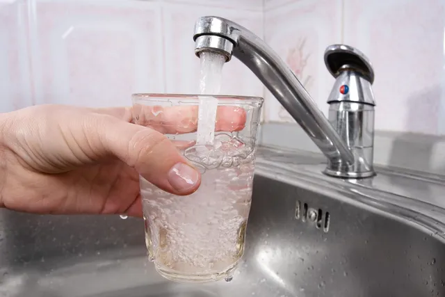 O mână ține un pahar de sticlă sub un robinet metalic din care curge apă într-o chiuvetă de inox.