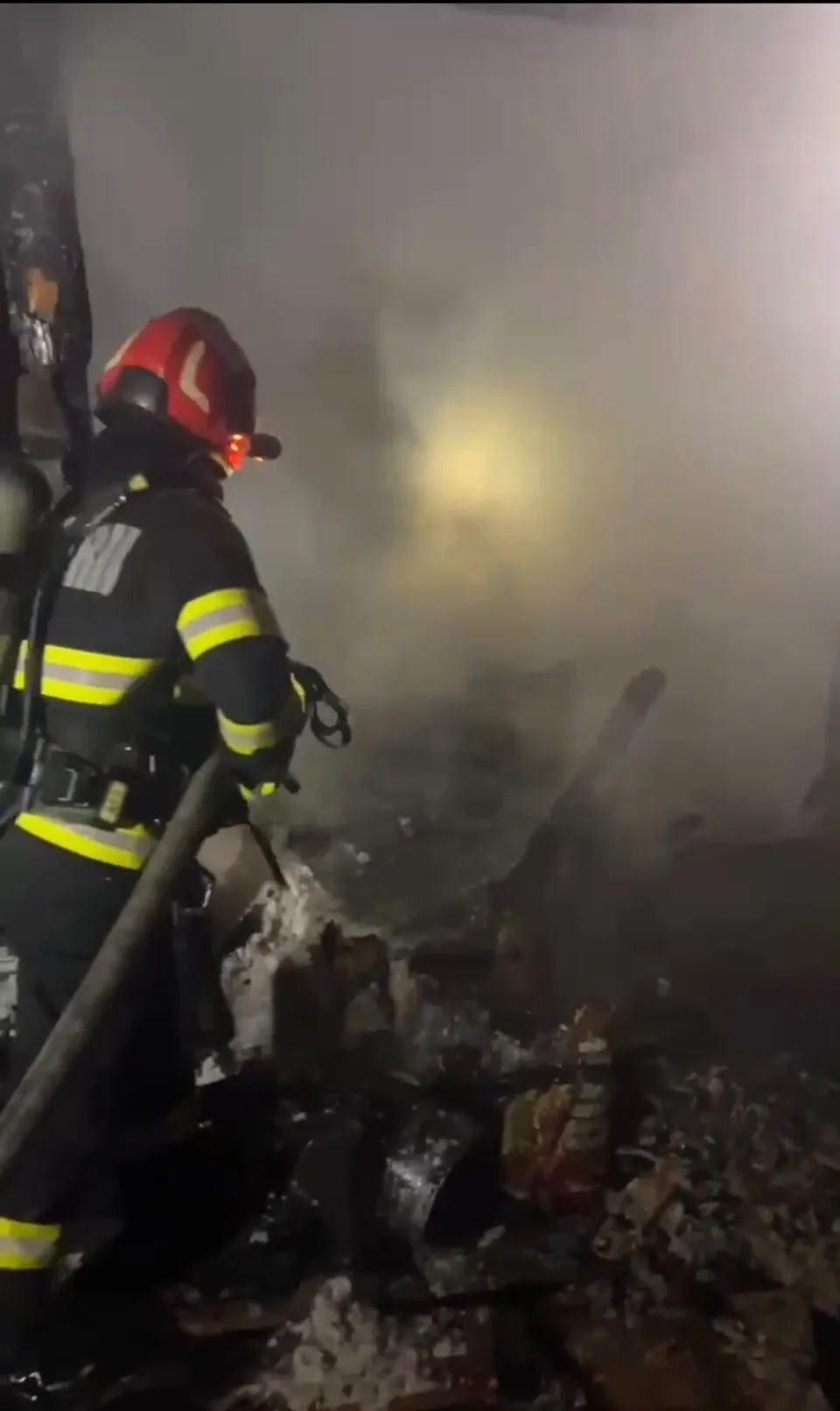 Un pompier cu echipament de protecție și cască este poziționat lângă un deșeu de materiale arse și fum dens într-un cadru nocturn.