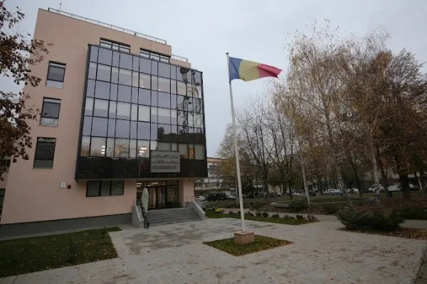 O clădire cu fațadă de sticlă și o steag românesc agitat pe un stâlp se află lângă un grup de copaci fără frunze.