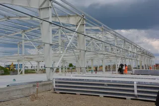 O hală cu structură metalică, cu stâlpi, grinzi, panouri ondulate și utilaje în fundal, sub cer noros.