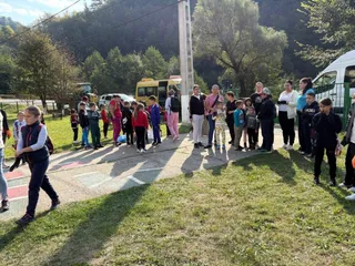 Un grup de copii și adulți stau pe o suprafață pavată lângă o zonă cu iarbă și copaci sub un cer luminos.