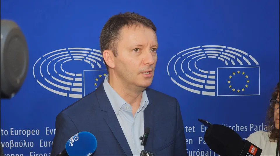 Un bărbat cu părul scurt și sacou albastru stă în fața unui fundal albastru cu emblemele Parlamentului European și microfoane în prim-plan.