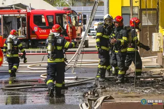 Cinci pompieri în echipament complet de protecție și cu echipamente de respirație stau lângă un autospecială de stingere și furtunuri pe sol.