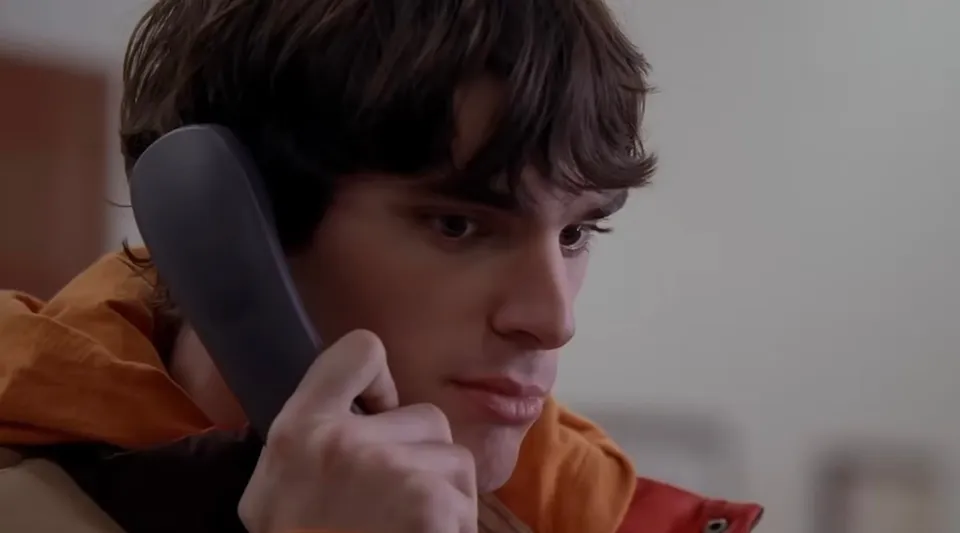 Un bărbat cu părul brun și o jachetă portocalie ține un telefon negru la ureche.