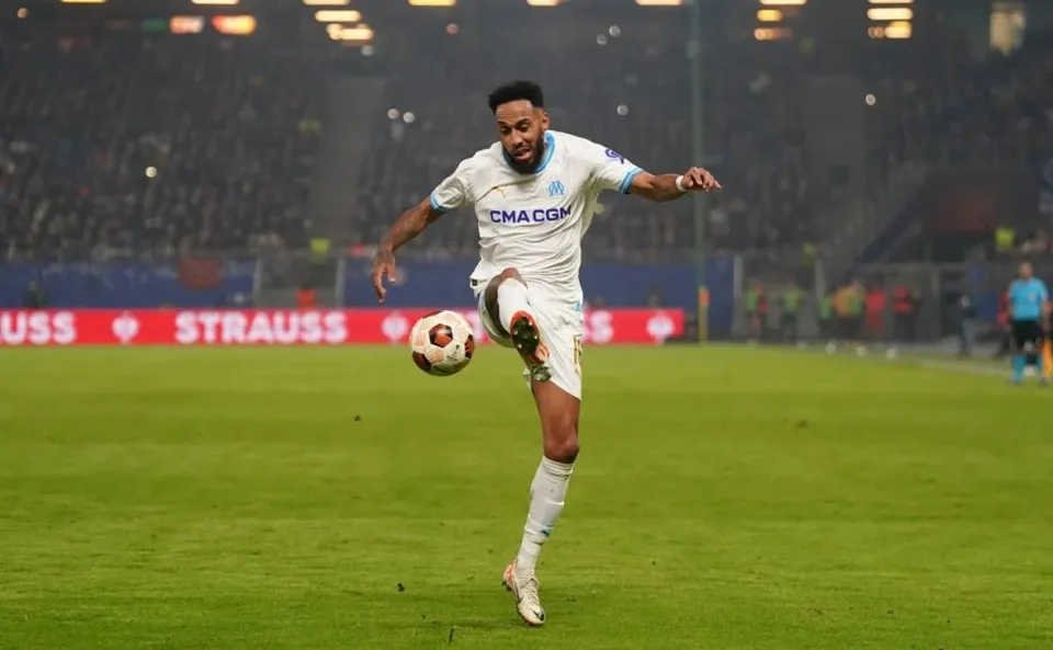 Un fotbalist într-un echipament alb de joc lovește un balon pe un teren de fotbal sub luminile unui stadion.