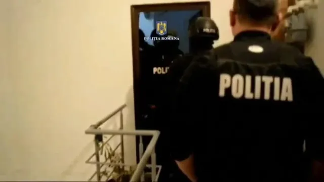 Un agent de poliție cu inscripția POLITIA pe spate privește spre o ușă deschisă prin care se văd alți doi agenți în uniformă întunecată.