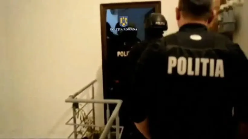Un agent de poliție cu inscripția POLITIA pe spate privește spre o ușă deschisă prin care se văd alți doi agenți în uniformă întunecată.
