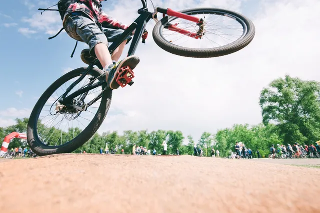 O persoană într-un tricou roșu și pantaloni scurți gri execută un salt cu o bicicletă neagră deasupra unui teren de pământ sub un cer senin.