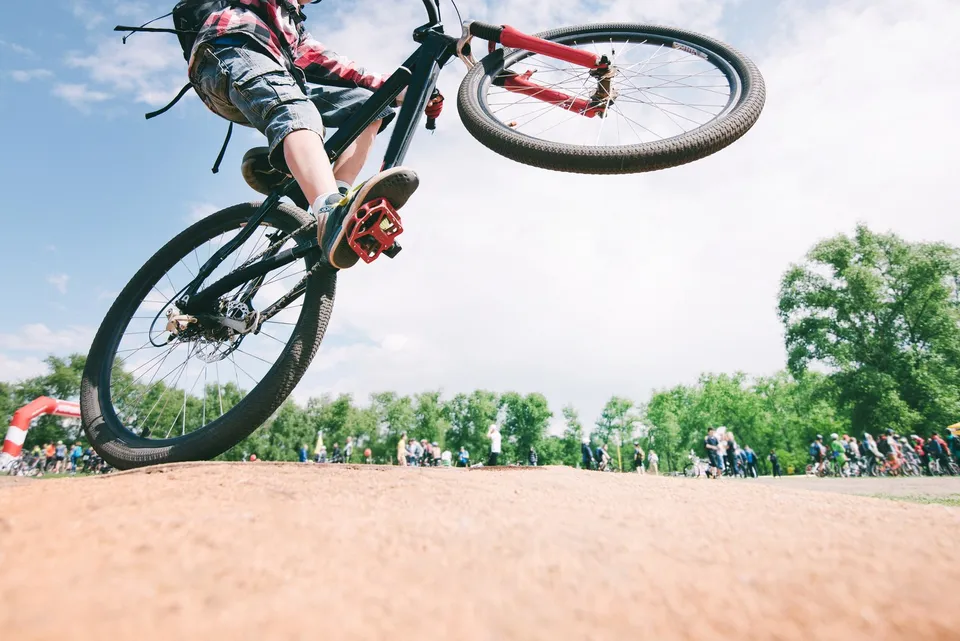 O persoană într-un tricou roșu și pantaloni scurți gri execută un salt cu o bicicletă neagră deasupra unui teren de pământ sub un cer senin.