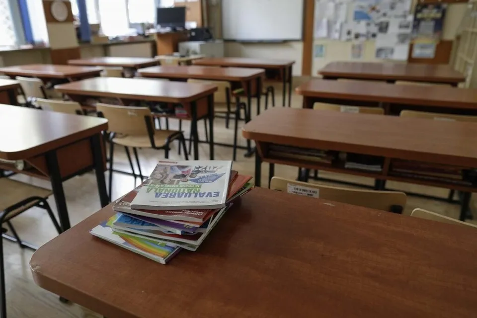 Mai multe birouri de lemn cu scaune din metal și câteva cărți colorate sunt așezate într-o sală de clasă.