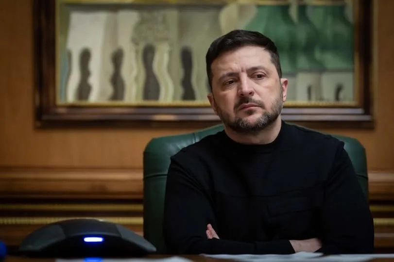 Un bărbat cu barbă și păr scurt, îmbrăcat într-un tricou negru, stă cu brațele încrucișate la o masă în fața unui tablou înrămat.
