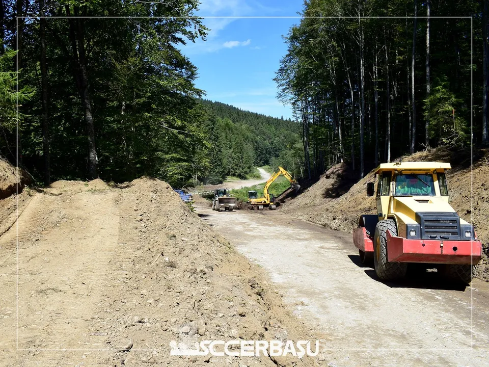 O excavatoare galbenă lucrează pe un drum de pământ lățit, lângă un utilaj de construcții galben și roșu situat în prim-plan.