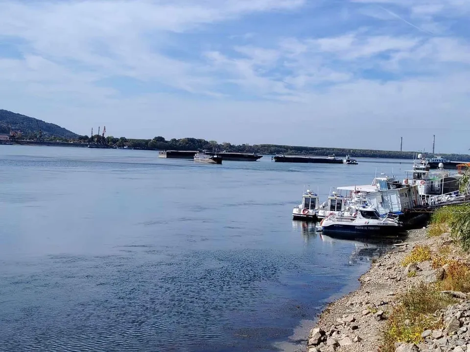 O barcă albă cu detalii albastre este ancorată lângă un mal pietros, în timp ce alte nave navighează pe un fluviu sub un cer înnorat.