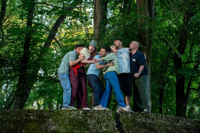 Un grup de șase persoane stă pe un zid de piatră într-o pădure, în timp ce unii dintre ei se îmbrățișează sau se țin de îmbrăcăminte.
