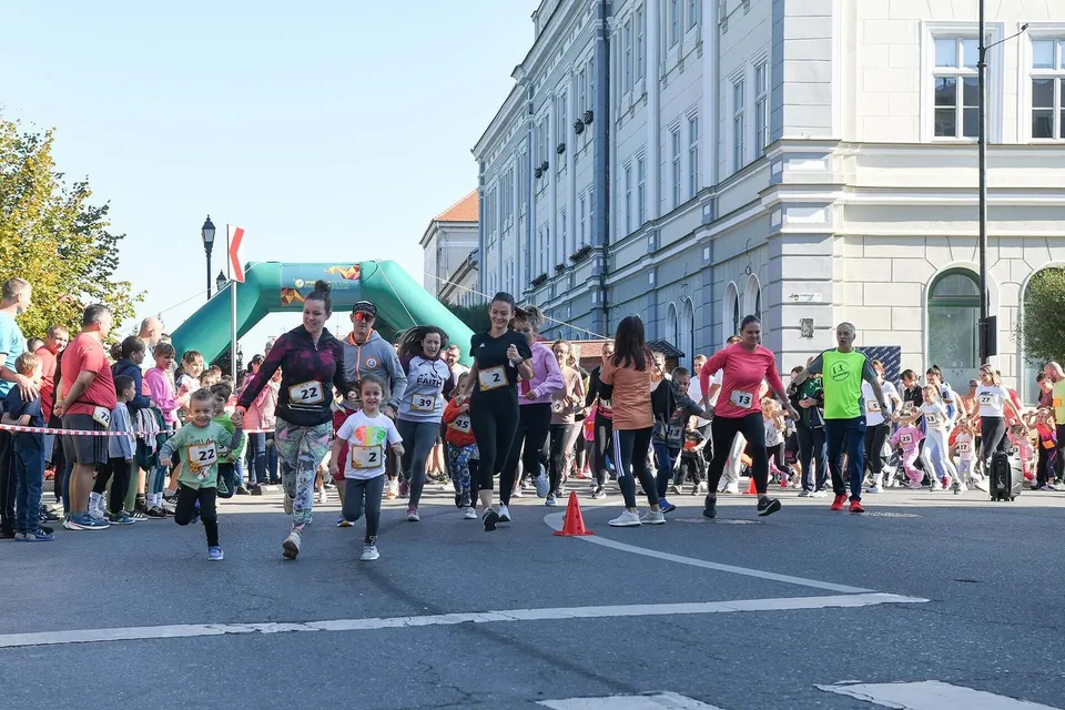 Oamenii aleargă pe un drum asfaltat prin fața unei clădiri mari, fiind încadrați de un arc gonflabil și de spectatori pe marginea drumului.