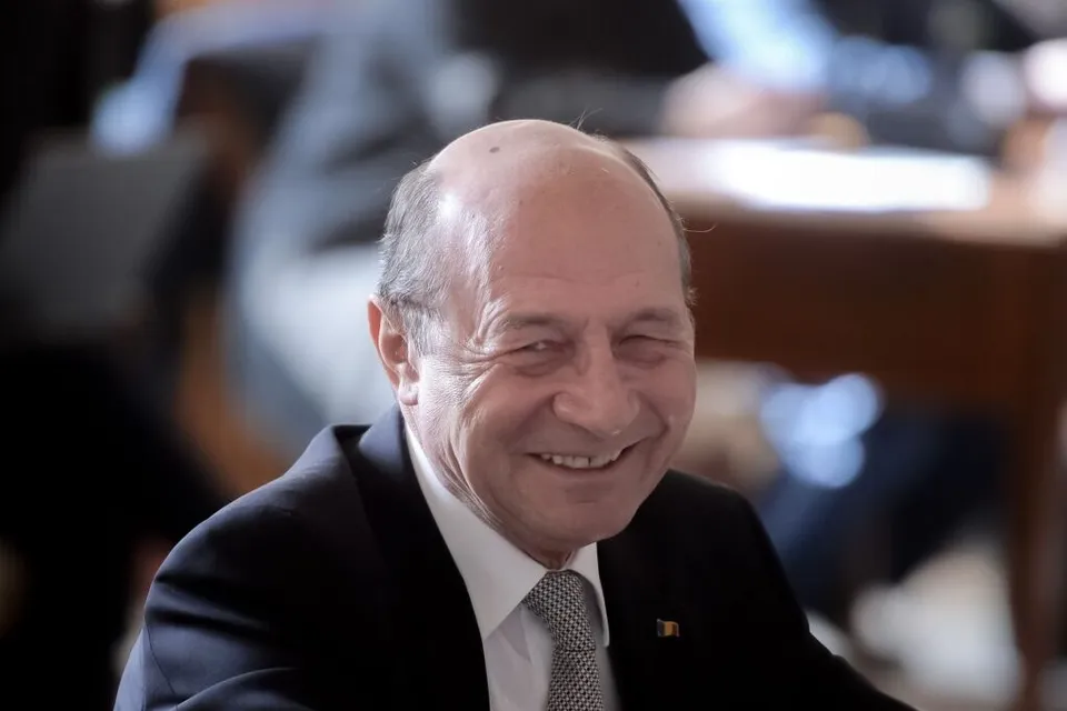 Un bărbat cu părul rare, purtând costum negru, cămașă albă și cravată gri, zâmbește într-un decor cu fundal neclar.