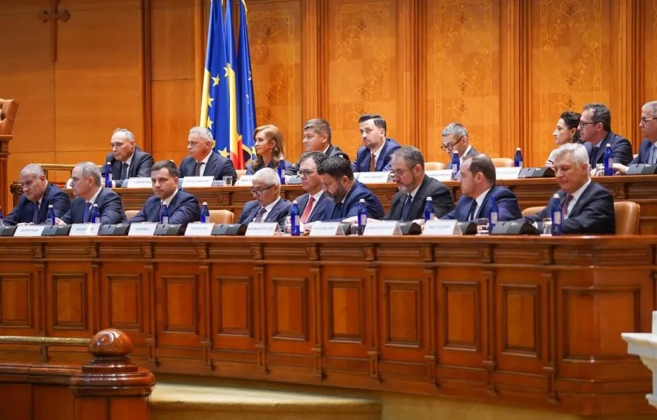 Un grup de bărbați în costume oficiale stau așezați la o masă lungă de lemn în fața unui panou de lemn și a unor steaguri.