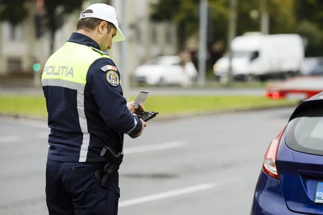 Un polițist cu uniformă albastră și galben deschide un dispozitiv electronic pe marginea unei străzi lângă o mașină albastră.