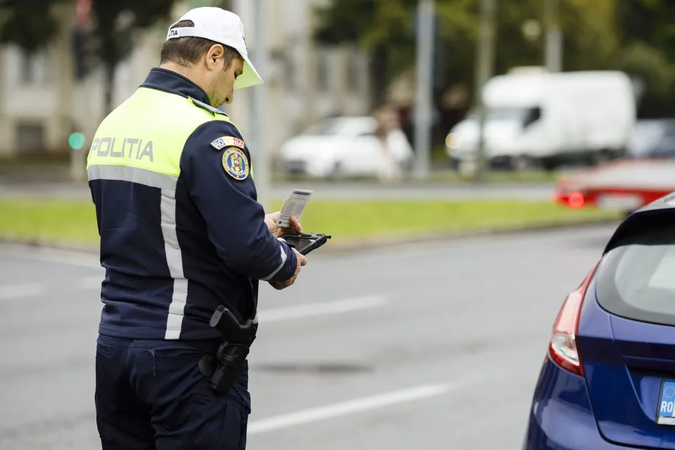 Un polițist cu uniformă albastră și galben deschide un dispozitiv electronic pe marginea unei străzi lângă o mașină albastră.