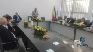 Oamenii stau așezați la mese lungi într-o încăpere cu pereți albi, având pe mese flori și obiecte mici.