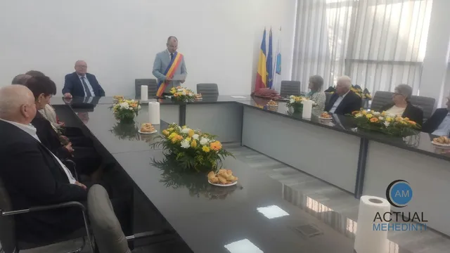 Oamenii stau așezați la mese lungi într-o încăpere cu pereți albi, având pe mese flori și obiecte mici.