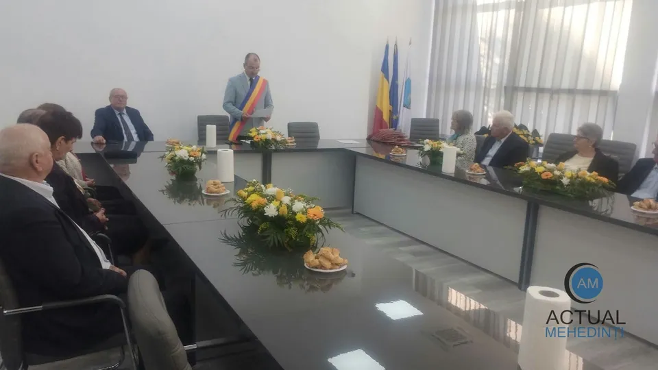 Oamenii stau așezați la mese lungi într-o încăpere cu pereți albi, având pe mese flori și obiecte mici.