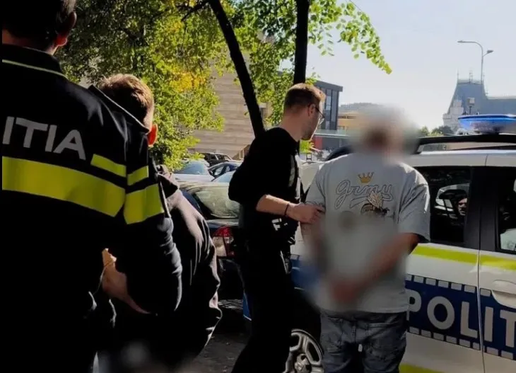 Un polițist în uniformă întunecată stă lângă un vehicul de poliție lângă un bărbat cu tricou alb și pantaloni gri.