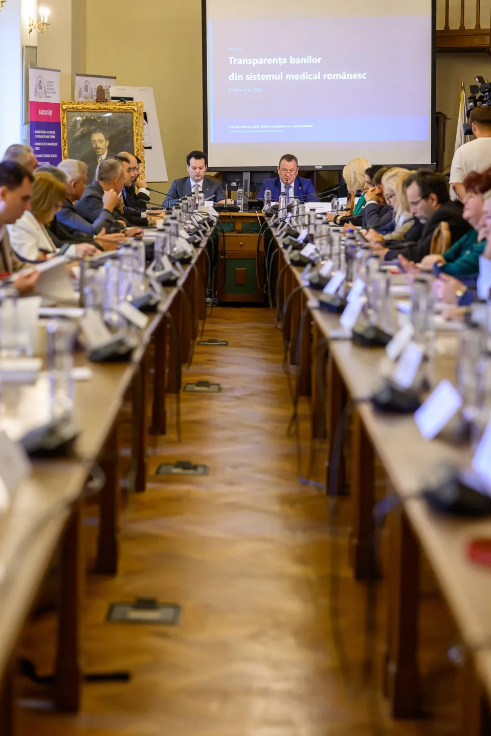 Oamenii stau la mese lungi de conferință într-o sală, privind spre un ecran de proiecție aflat în partea din spate a încăperii.