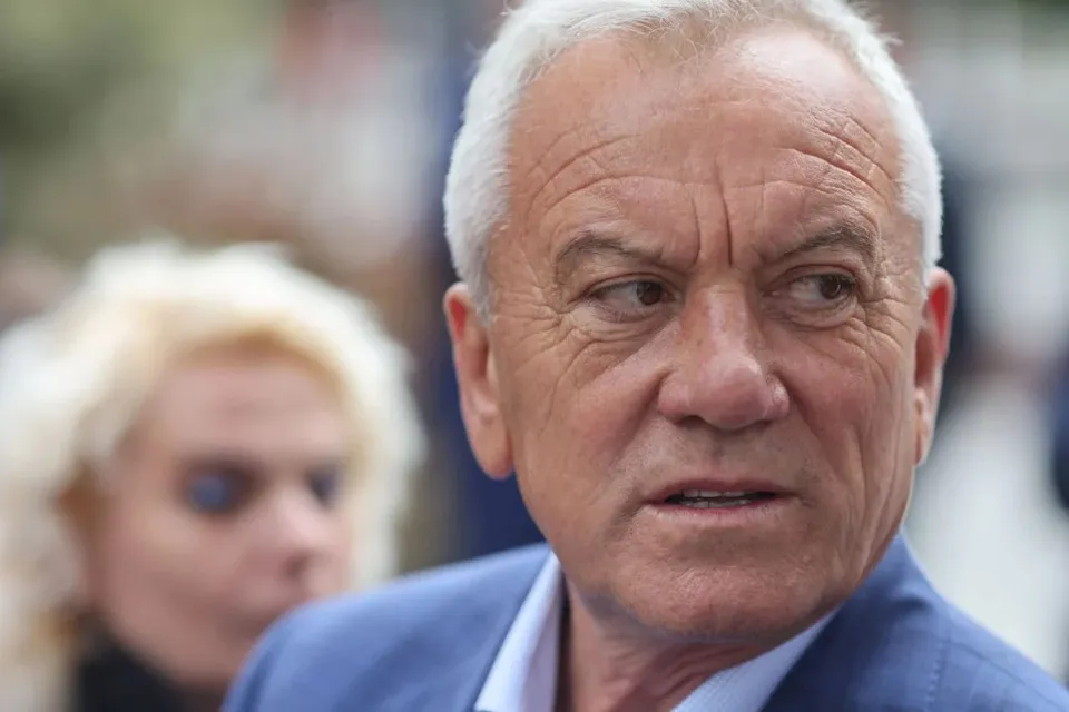 Un bărbat cu părul scurt și alb gri poartă un sacou albastru și o cămașă deschisă, privind spre lateral.
