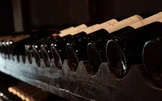 Mai multe sticle de vin negre sunt depozitate într-un suport de lemn întins pe orizontală într-o încăpere întunecată.