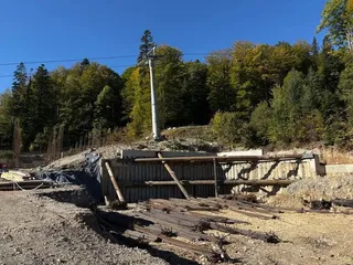 Un pilon metalic înalt stă pe un teren accidentat lângă o fundație de beton și scânduri de lemn într-o zonă împădurită.