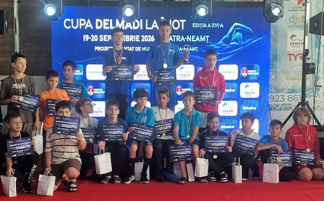 Un grup de copii stă în rând pe un podium roșu, purtând medalii și ținând diplome sub un panou albastru cu textul ediției a XVI-a Cupei Delmadi.