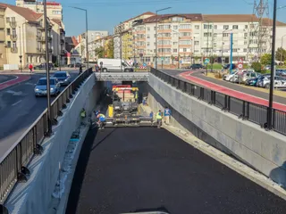 Mașini de asfaltare și lucrători cu veste reflectorizante execută lucrări pe o porțiune de drum proaspăt asfaltat într-un pasaj subteran cu clădiri în fundal.