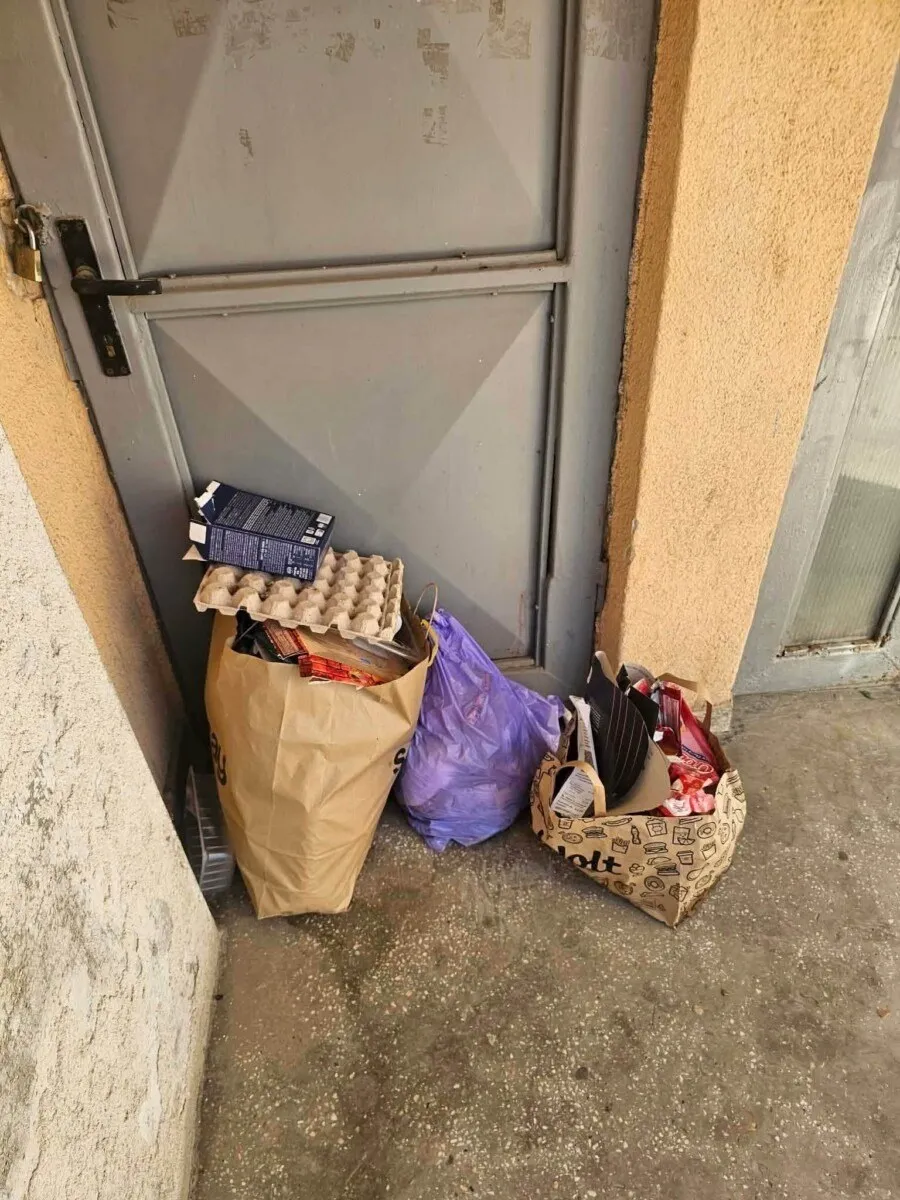 Mai multe saci de gunoi și cutii de carton sunt depozitate pe sol în fața unei uși metalice gri.