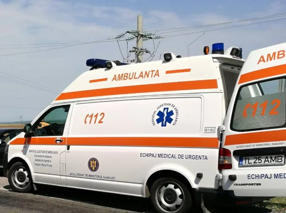 Două ambulanțe albe cu benzi portocalii, inscriptionate cu „AMBULANTA” și „112”, sunt parcate lângă una dintre ele, având vizibil și semnalizatorul optic albastru.