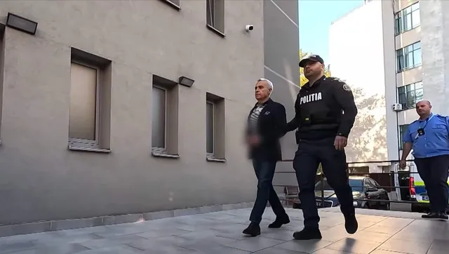 Un bărbat în costum și un polițist în uniformă neagră merg pe o suprafață pavată lângă o clădire cu ferestre înguste.