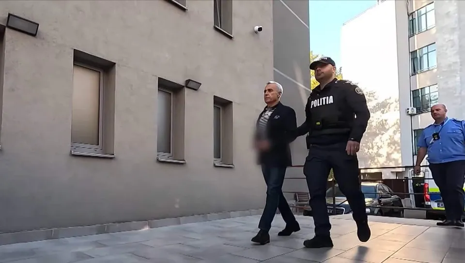 Un bărbat în costum și un polițist în uniformă neagră merg pe o suprafață pavată lângă o clădire cu ferestre înguste.
