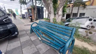 Un banc de grădină de culoare albastră este situat pe un trotuar de pavaj lângă un microcar alb și un vehicul negru.