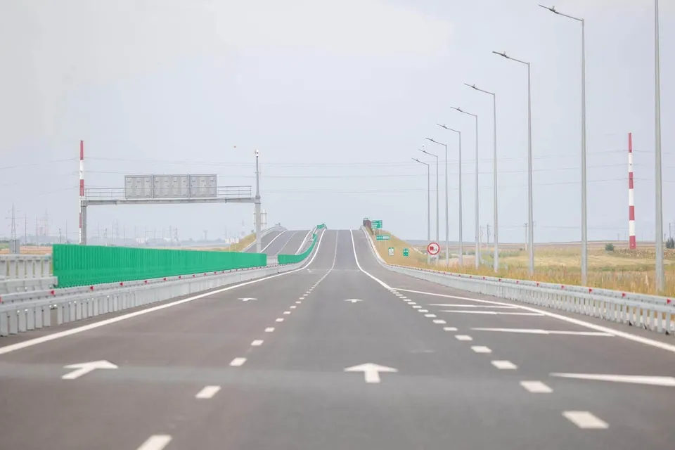 O autostradă cu mai multe benzi de circulație, marcaje albe pe asfalt și stâlpi de iluminat dispuiși pe partea dreaptă sub un cer înnorat.